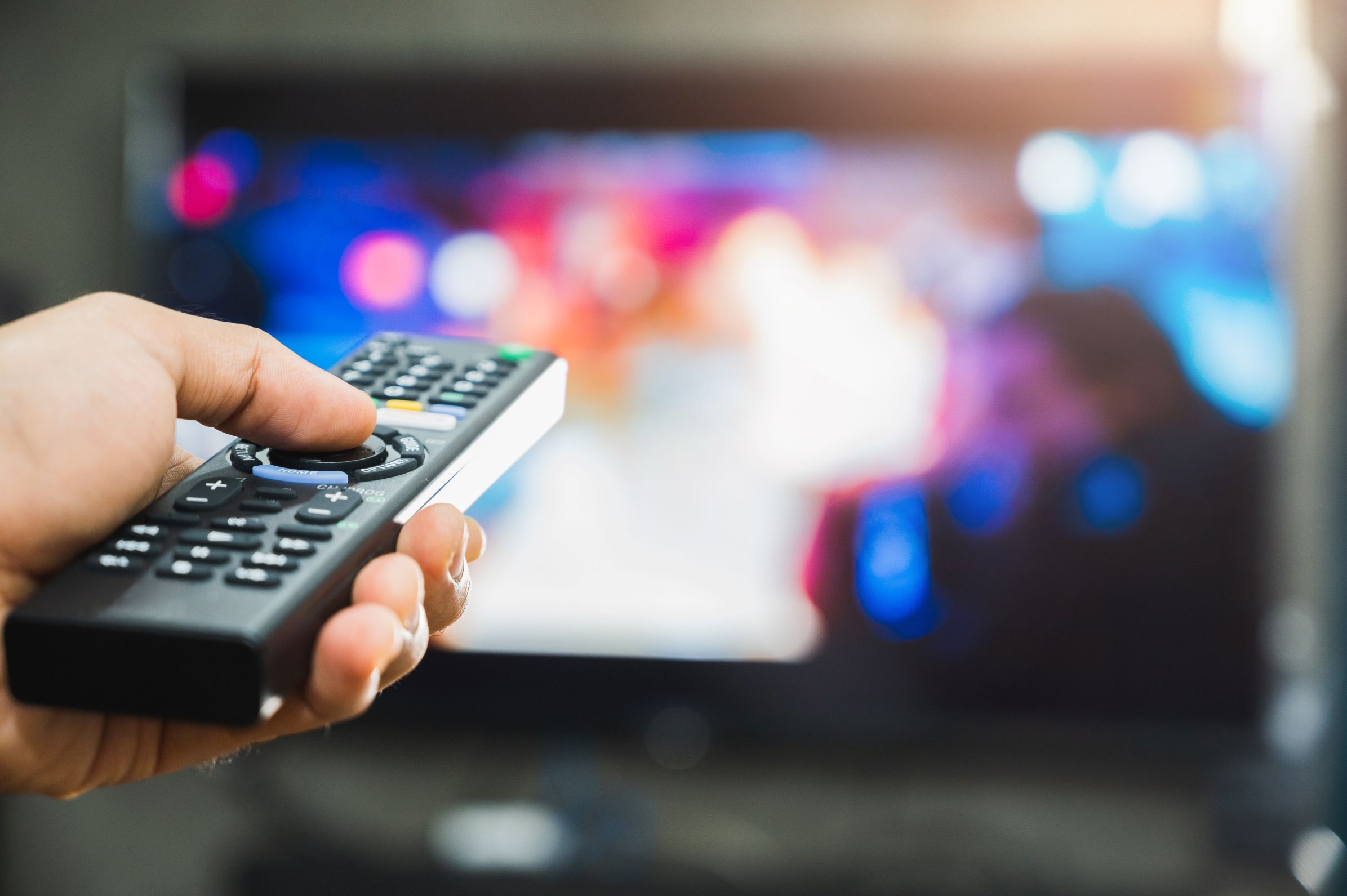 Closeup of a man's hand pointing a remote control at a TV in the background.